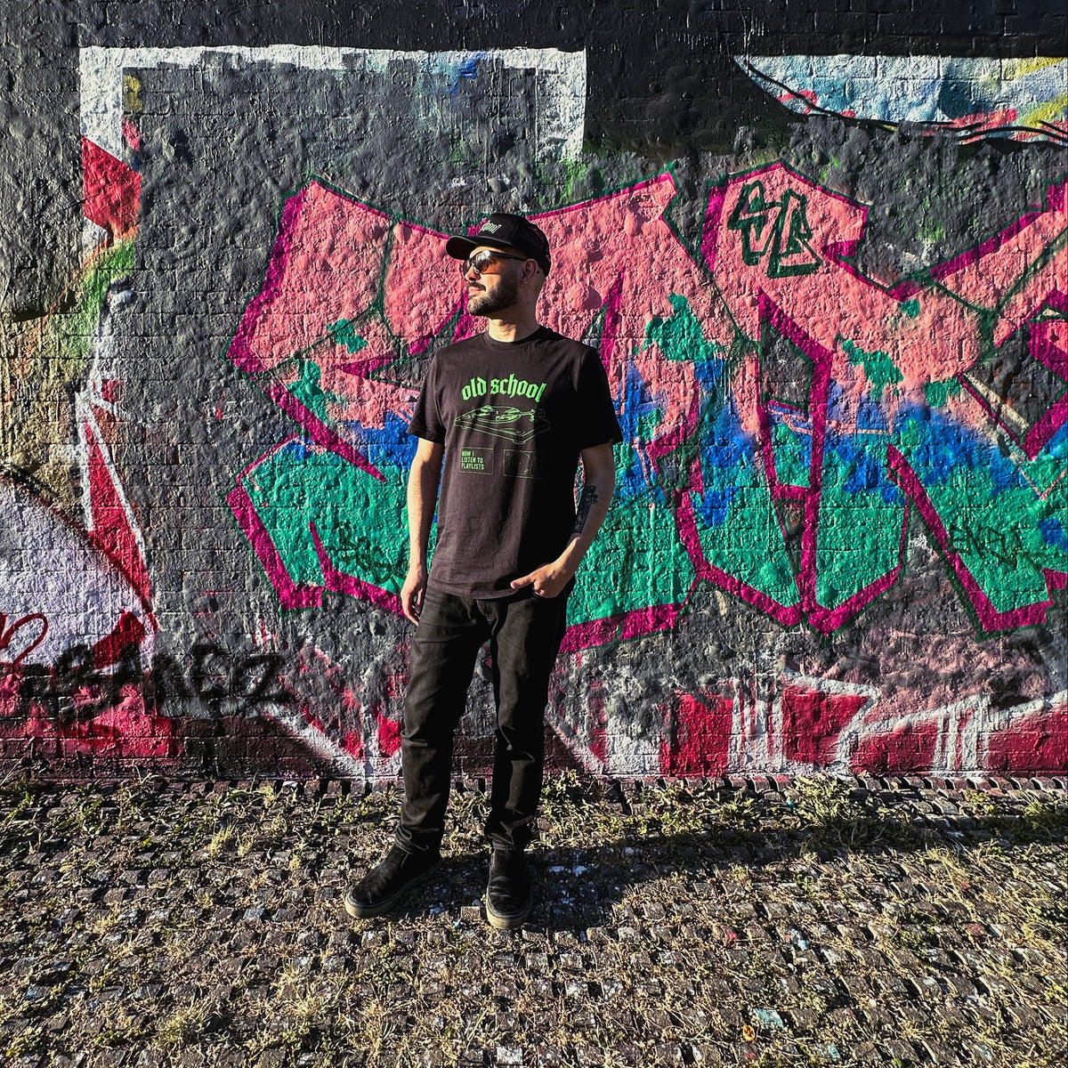 Young Brazilian Man wearing a Tropical Fuzz fairwear organic vegan cotton unisex black t-shirt with 'old school' type and a vinyl record player in bright neon green design, in front of a colourful graffiti wall.
