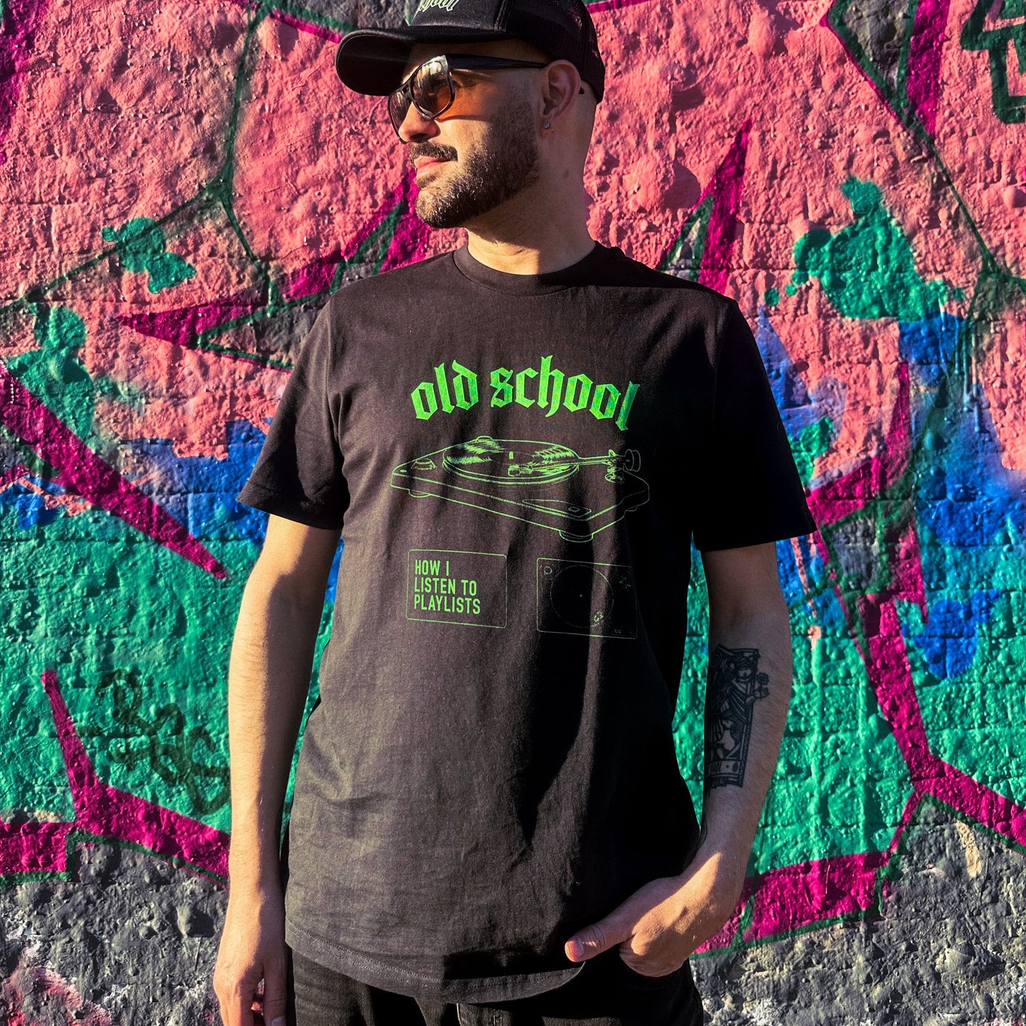 Man wearing a black t-shirt with 'old school' text in green, standing in front of a colorful graffiti wall.