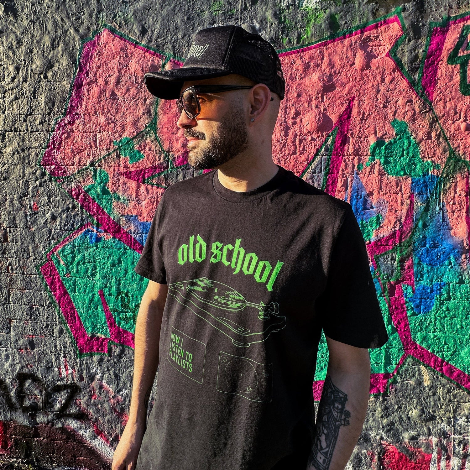 Young Brazilian Man wearing a Tropical Fuzz fairwear organic vegan cotton unisex black t-shirt with 'old school' type and a vinyl record player in bright neon green design, in front of a colourful graffiti wall.