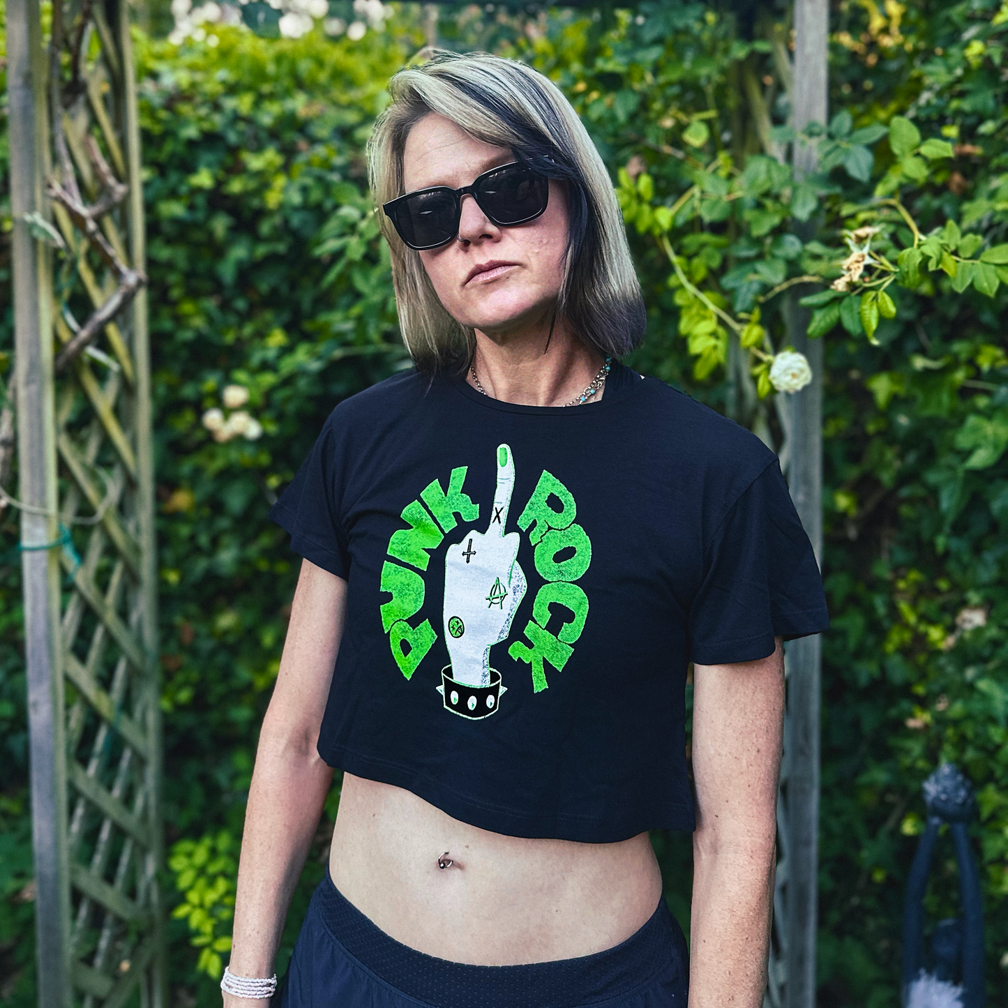 A woman wearing a black cropped t-shirt with a green and white 'Dunk Rock' graphic, standing outdoors in front of a green garden trellis.