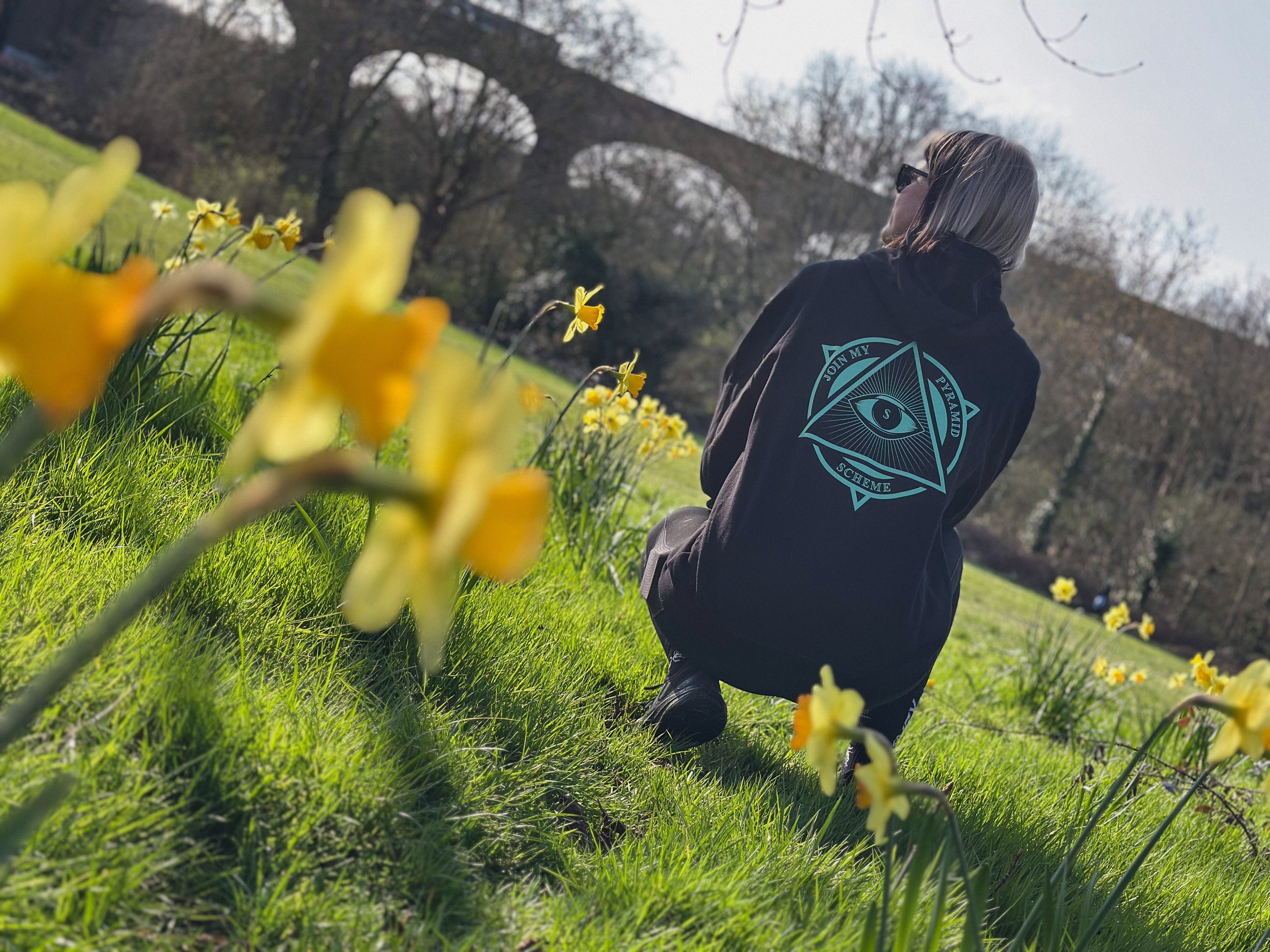 Person wearing a black hoodie with a symbol on the back, standing in a field of yellow flowers with a bridge in the background.