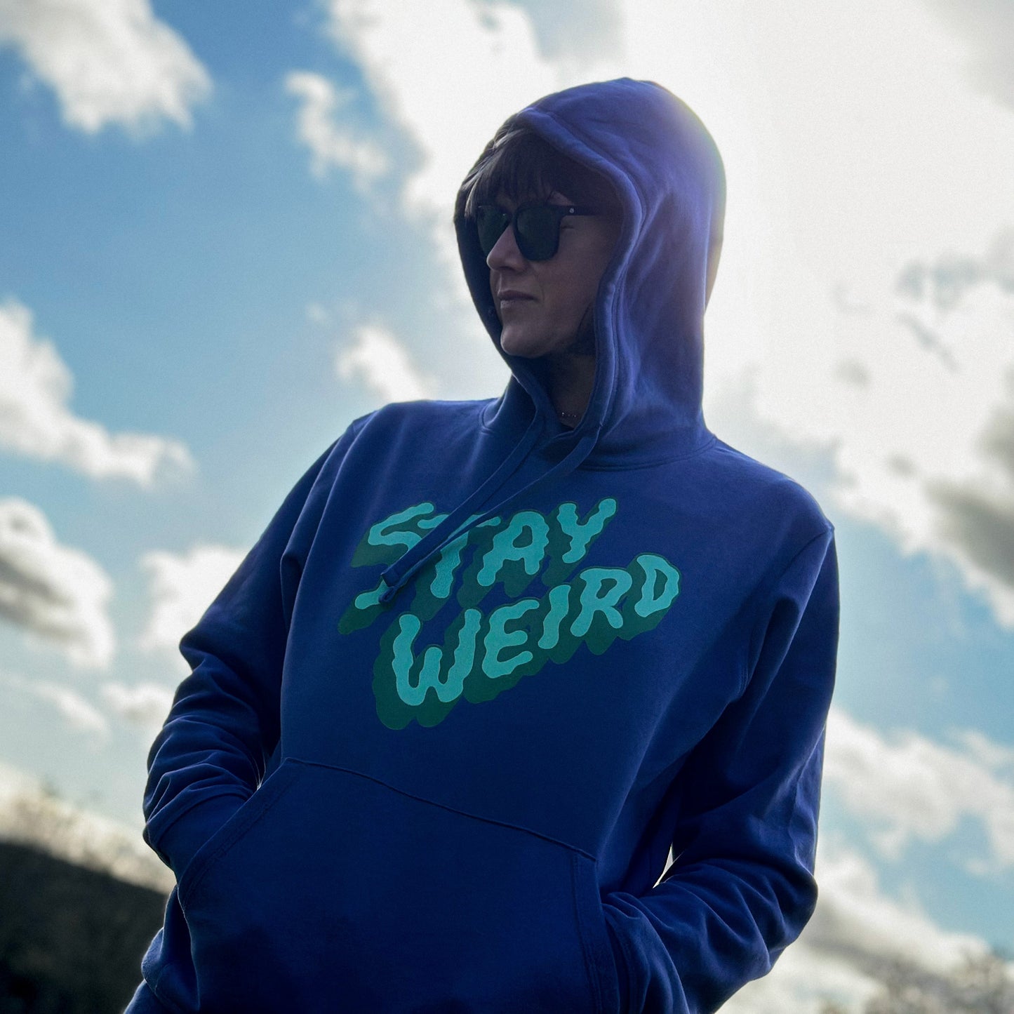 Person wearing a purple hoodie with 'Stay Weird' text against a cloudy sky.