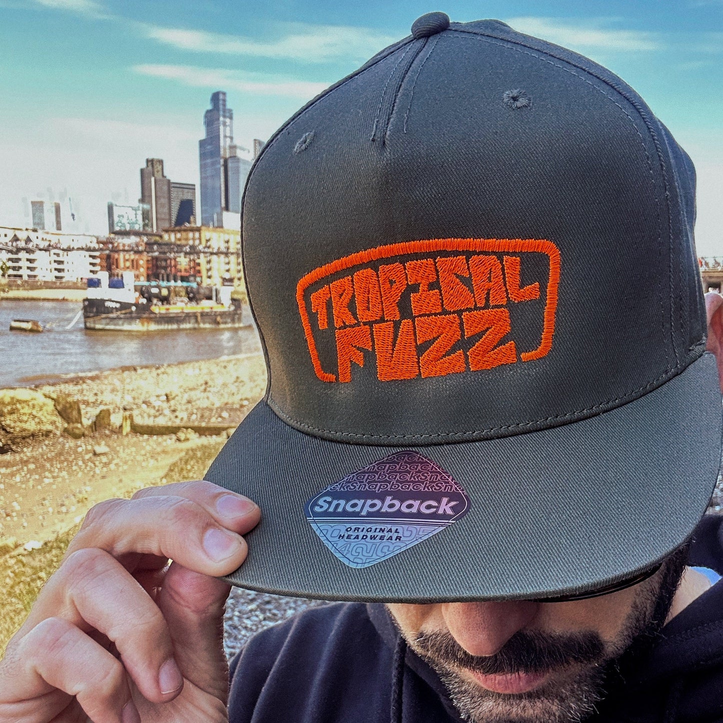 Person wearing a gray cap with 'Tropical Fuzz' logo, against the London urban skyline.