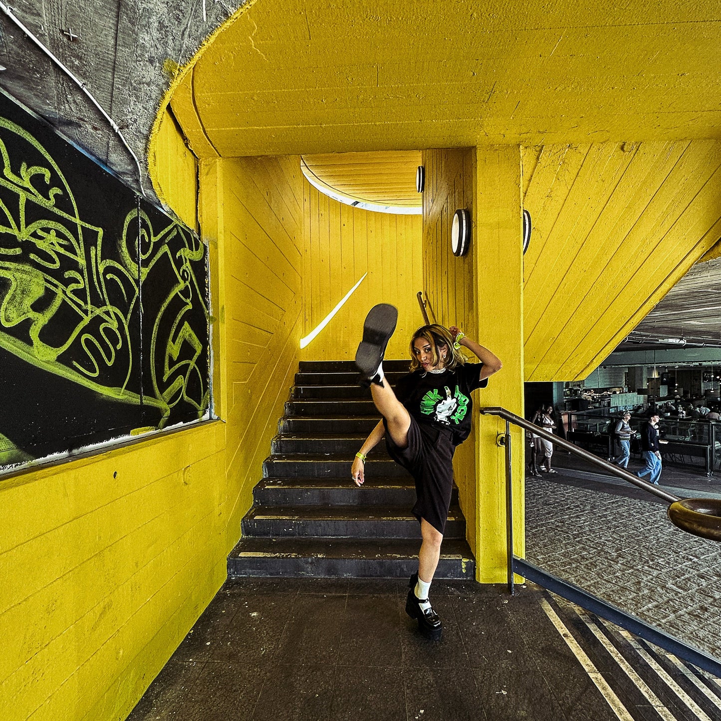 Person wearing black punk rock tshirt in an urban setting with yellow walls and stairs.