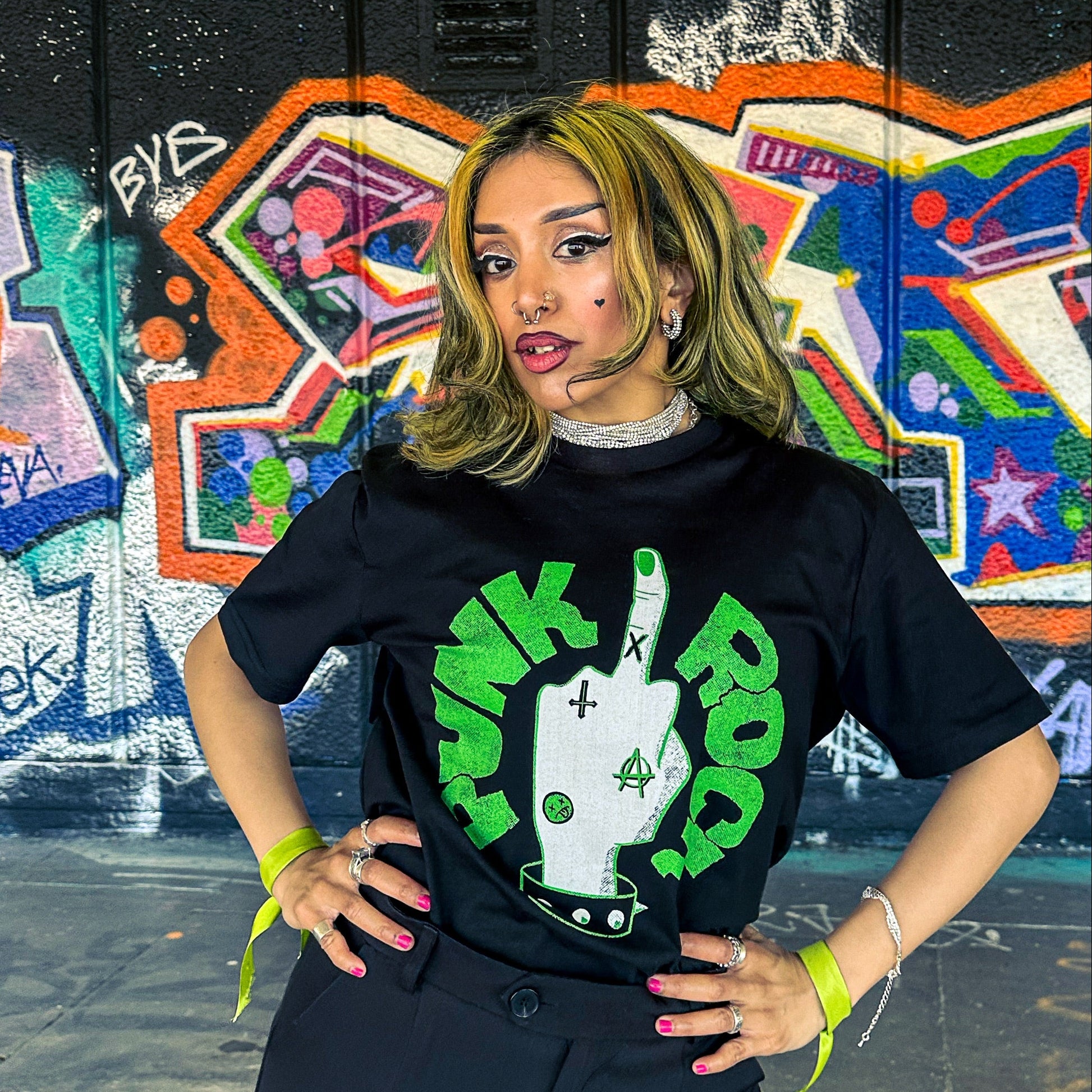 Person wearing a black t-shirt with green and white graphic design in front of a colorful graffiti wall.