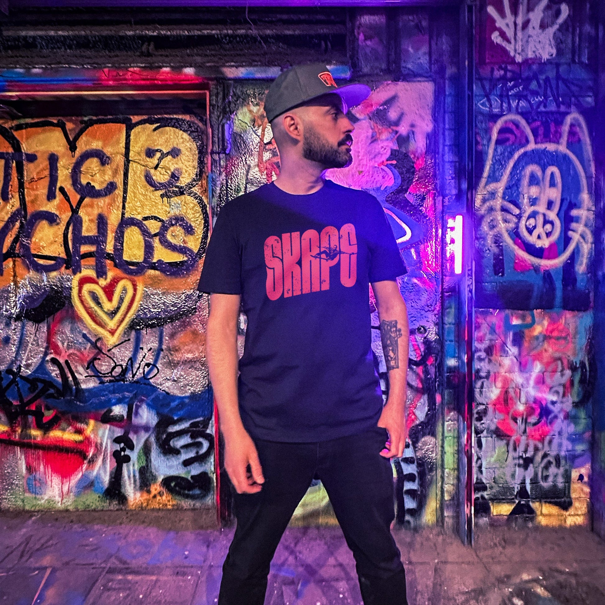 Person wearing a dark t-shirt with visible text standing in front of a graffiti-covered wall at night.