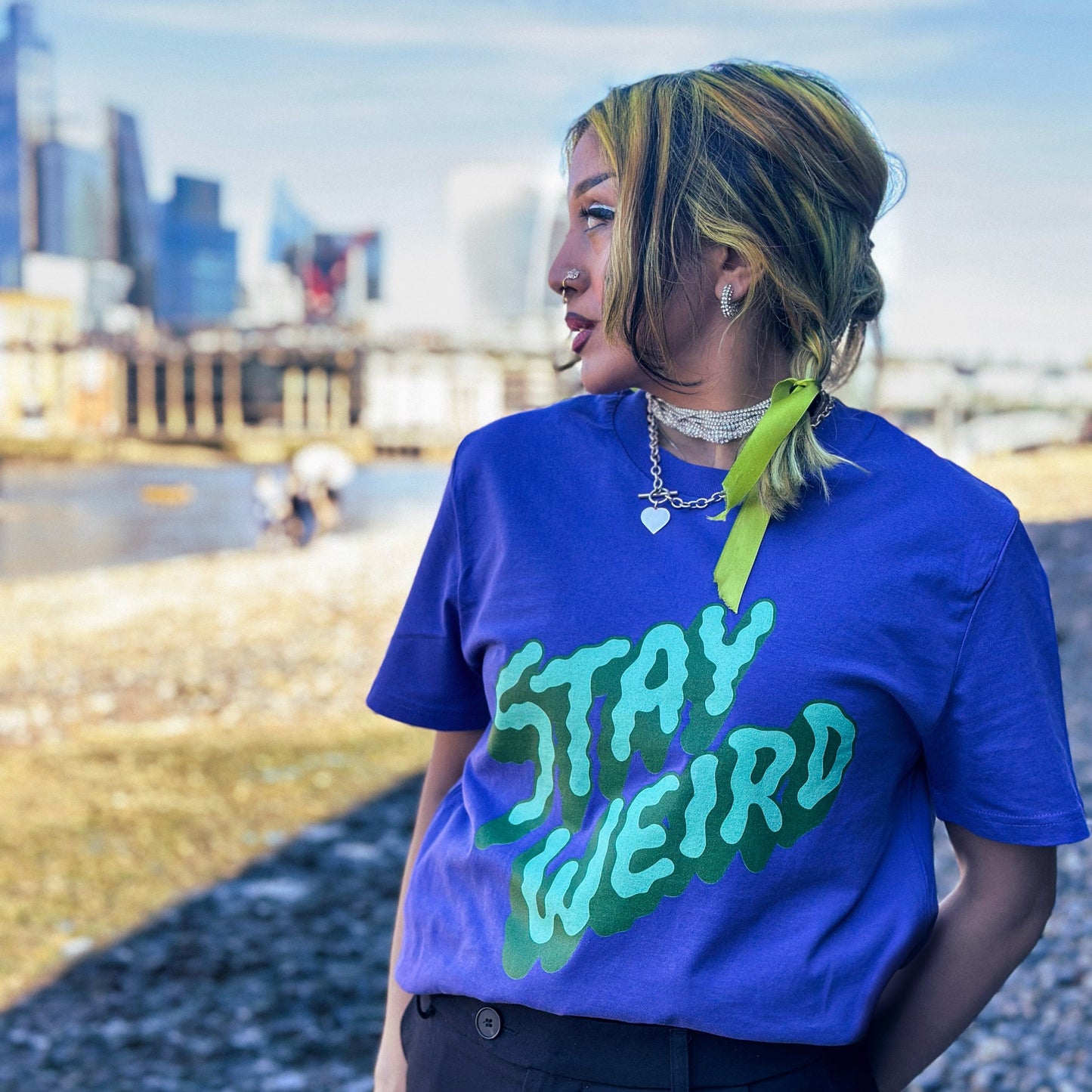 Person wearing a purple t-shirt with 'Stay Weird' text, standing by the Thames river with the London skyline in the background.