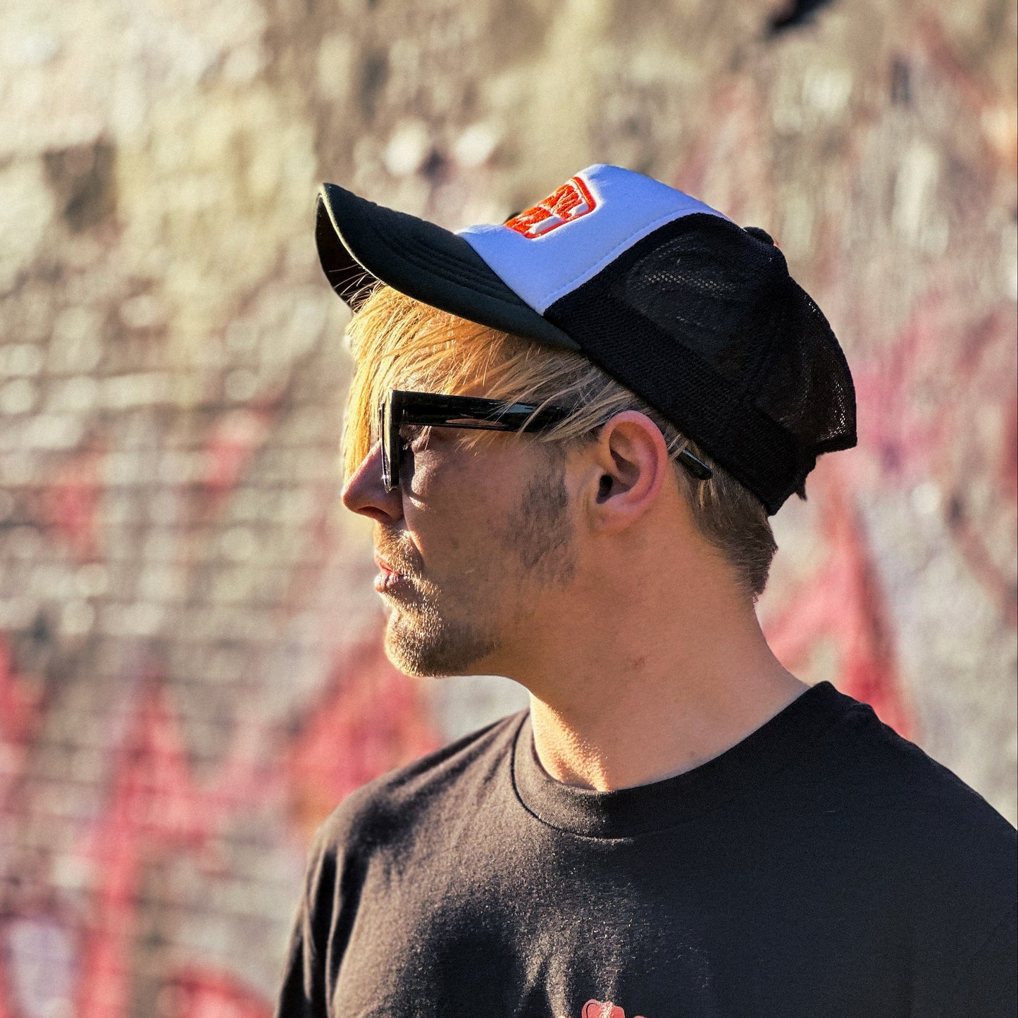 Person wearing a black 'Tropical Fuzz' t-shirt with a blue and black cap in front of a graffiti-covered wall.