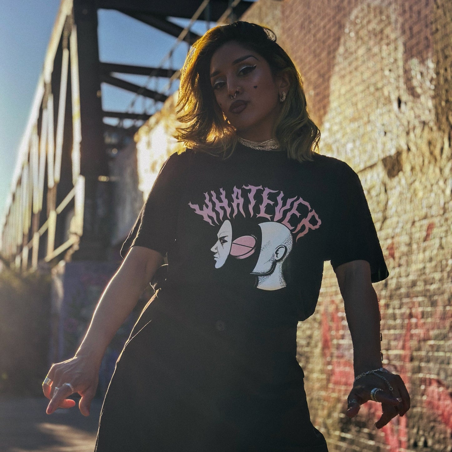 Person wearing a black t-shirt with graphic design and text, standing in front of a bridge and wall with graffiti.