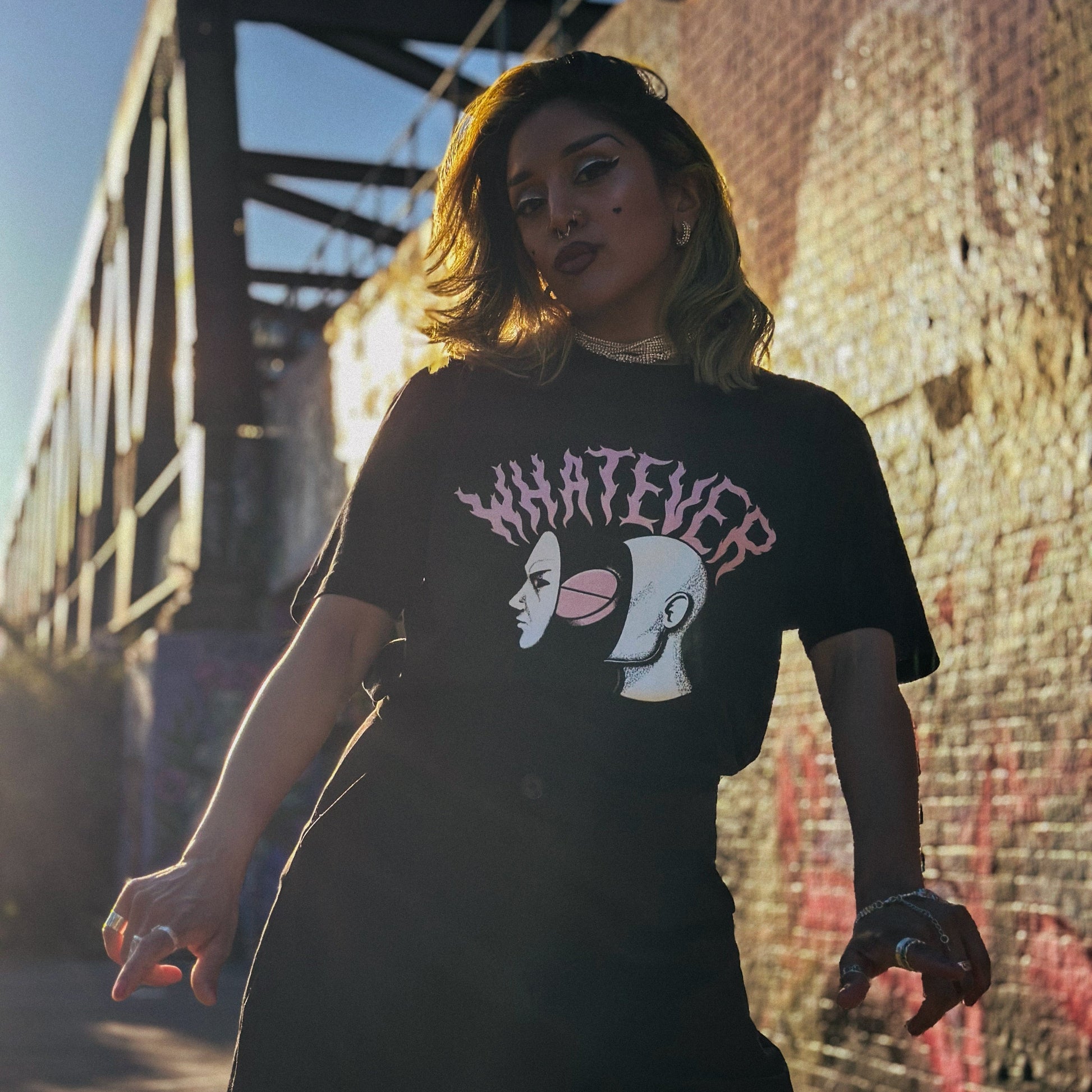 Person wearing a black t-shirt with graphic design and text, standing in front of a bridge and wall with graffiti.