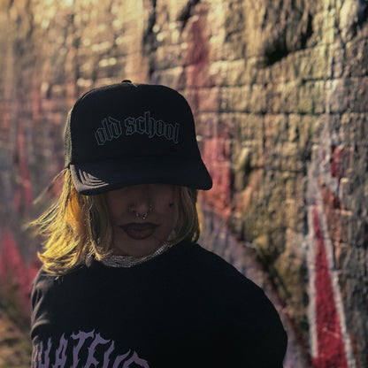 Person wearing a black cap with the text 'old school', standing against an urban graffiti-covered wall.