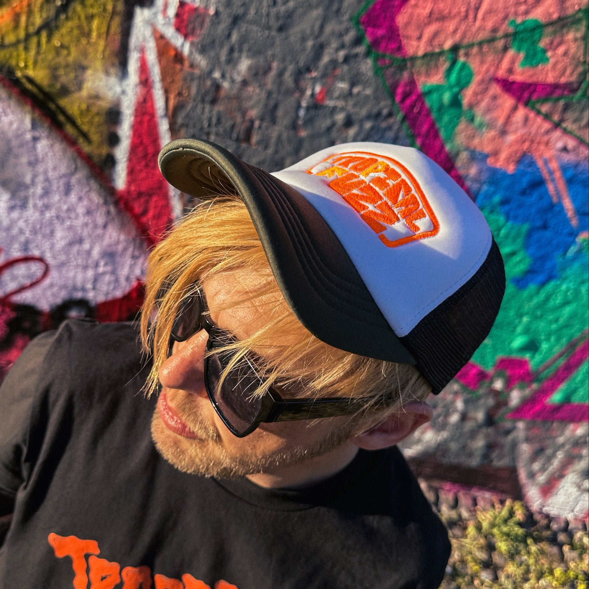 Person wearing a cap with a logo and a black t-shirt with orange text, standing in front of colorful graffiti.